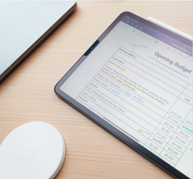 Tablet displaying an opening budget spreadsheet with handwritten notes, placed on a wooden desk beside a laptop and wireless charger.